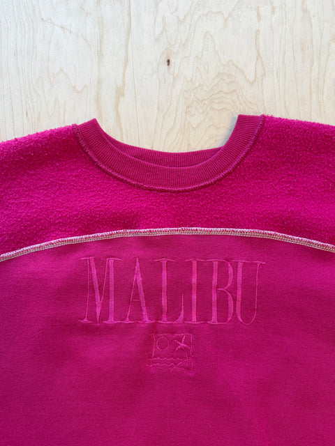 Pink fleece pullover with 'Malibu' branding on a wooden surface